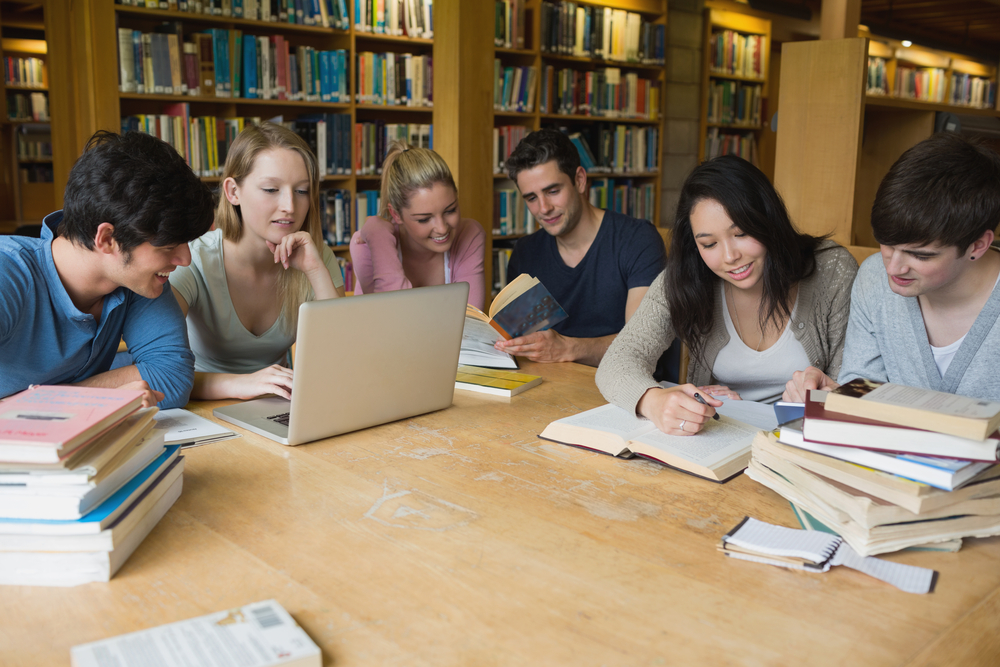 Students sitting at a table in a library while learning and working on a laptop