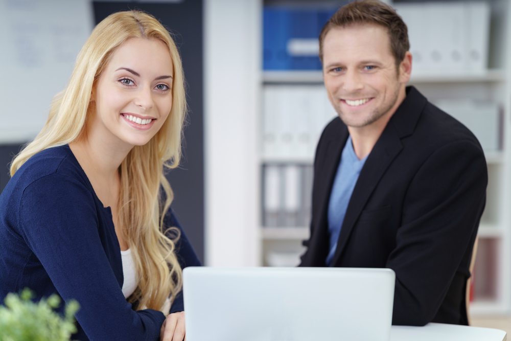 Successful young business couple working together on a self-employed business posing at a desk in their office smiling at the camera
