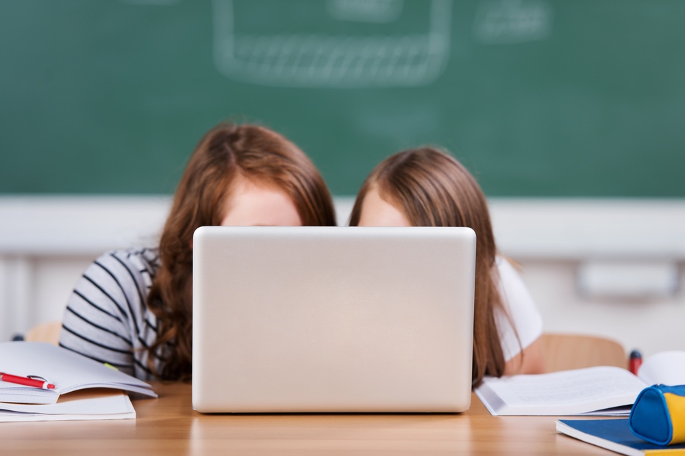 Two schoolgirls hiding behind the laptop in the classroom