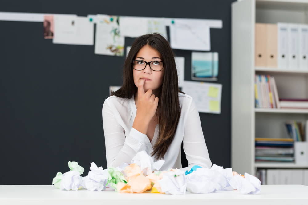 Young woman with writers block sitting in an office with a desk littered with crumpled paper as she sits looking thoughtfully into the air with her finger to her chin seeking new ideas-1